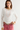 A woman modeling a white ribbed long-sleeve top with lace trim at the neckline and lace panels on the sleeves, paired with pink jeans and layered necklaces, against a plain light gray studio background.