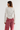 A woman shown from the back wearing a white ribbed long-sleeve top with lace trim at the neckline and lace cuffs, paired with dusty rose jeans, against a plain light gray studio background.