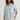 A woman wearing a light blue and white vertically striped button-down shirt with rolled sleeves and cream-colored pants, photographed against a plain light gray studio background.