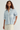 A woman wearing a light blue and white vertically striped button-down shirt with rolled sleeves and cream-colored pants, photographed against a plain light gray studio background.
