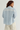 A woman shown from the back wearing a light blue and white vertically striped button-up shirt and cream-colored pants, photographed against a plain light gray studio background.
