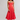 A woman modeling a coral red midi dress with a sweetheart neckline, ruffled straps, and a ruffled hem, paired with brown leather sandals against a light gray studio background.