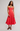 A woman modeling a coral red midi dress with a sweetheart neckline, ruffled straps, and a ruffled hem, paired with brown leather sandals against a light gray studio background.