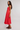 A woman modeling a bright coral-red midi dress with thin straps and a ruffled hem, paired with dark brown flat sandals, against a plain light gray studio background.