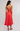 A woman wearing a coral red sleeveless midi dress with thin straps, a ruffled hem, and a low open back, photographed from behind against a light gray studio background.