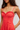 A close-up of a model wearing a coral red spaghetti-strap dress with a sweetheart neckline and structured bodice against a light gray background.