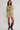 A woman modeling a light olive-beige knit mini dress with a lace-up keyhole neckline, paired with dark brown knee-high leather boots, against a plain light gray studio background.