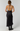 A woman shown from the back wearing a black satin halter slip dress with tie-back straps and black ankle boots, photographed against a clean white studio background.