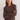 A woman with long light brown hair wearing a brown ribbed knit long-sleeve sweater and blue jeans, standing against a plain light gray background with her arms crossed.