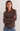A woman with long light brown hair wearing a brown ribbed knit long-sleeve sweater and blue jeans, standing against a plain light gray background with her arms crossed.