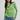 A woman wearing a lime green long-sleeve knit top with a frayed hem, paired with blue jeans and a chunky green beaded necklace, against a light gray studio background.