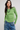 A woman wearing a lime green long-sleeve knit top with a frayed hem, paired with blue jeans and a chunky green beaded necklace, against a light gray studio background.