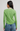 A woman viewed from the back wearing a light green long-sleeve knit top with a scalloped hem and a patterned neck scarf, paired with blue jeans against a light gray studio background.