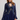 A woman modeling a navy blue lace long-sleeve top with a high neck and ruffled sleeves, paired with blue jeans, against a light gray studio background.