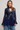 A woman modeling a navy blue lace long-sleeve top with a high neck and ruffled sleeves, paired with blue jeans, against a light gray studio background.