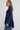 A woman modeling a navy blue lace long-sleeve top with a high mock neck and flared cuffs, paired with blue jeans, against a plain light gray background.