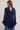 A woman shown from the back wearing a navy blue lace long-sleeve top with a keyhole cutout and a matching navy pleated skirt, paired with blue jeans against a light gray studio background.