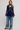 A woman modeling a navy blue lace long-sleeve top with a peplum hem and wide-leg blue jeans against a light gray studio background.