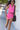 Woman in a pink outfit with a black bag standing against a white wall.