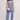 A woman modeling a light blue two-piece lounge set consisting of a sleeveless wrap-style crop top and matching jogger pants, styled with a lavender quilted clutch and beige slide sandals against a plain light gray studio background.