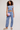 A woman modeling a light blue two-piece lounge set consisting of a sleeveless wrap-style crop top and matching jogger pants, styled with a lavender quilted clutch and beige slide sandals against a plain light gray studio background.