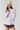 A model wearing a light lavender long-sleeve boatneck t-shirt, dark purple athletic shorts, and a cream-colored knit beanie hat, posed against a solid light gray studio background.