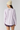 A model shown from the back wearing a light lavender long-sleeve crewneck sweatshirt, a cream knit beanie, and dark purple shorts against a light gray studio background.