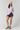 A model stands against a light gray studio background wearing a light purple long-sleeve top, dark purple athletic shorts, a cream knit beanie, gray socks, and beige hiking shoes.