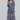 A woman modeling a long-sleeve ruffled maxi dress in a dusty slate blue-gray color, paired with dark brown Mary Jane flats, against a solid light gray studio background.