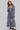A woman modeling a long-sleeve ruffled maxi dress in a dusty slate blue-gray color, paired with dark brown Mary Jane flats, against a solid light gray studio background.