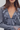 A close-up of a woman wearing a slate blue-gray ruffled blouse with a deep V-neckline and layered ruffles, styled with a silver teardrop pendant necklace.