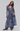 A model wearing a long, ruffled, slate blue-gray sheer cardigan or cover-up layered over wide-leg blue jeans, photographed against a neutral light gray studio background.