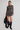 A model stands against a light gray studio background wearing a long-sleeve mini dress with a brown, blue, and red paisley pattern, paired with black knee-high boots and holding a black jacket.