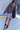 A woman from behind wearing an oversized taupe-brown hoodie, light lavender shorts, black socks, and dark loafers, carrying a black shoulder bag while walking on a concrete sidewalk.
