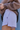A close-up lifestyle shot of a person wearing light lavender athletic shorts with a side slit and a small embroidered leaf logo, paired with a brown sweatshirt.