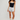 A model stands against a light gray studio background wearing a black camisole with a white band, light gray shorts, beige leg warmers, and black sneakers.