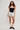 A model stands against a light gray studio background wearing a black camisole with a white band, light gray shorts, beige leg warmers, and black sneakers.
