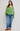 A woman modeling a light green long-sleeve blouse with a scoop neckline, gathered sleeves, and tie details at the cuffs, paired with blue jeans and brown suede espadrilles against a light gray studio background.