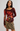 A woman modeling a long-sleeve scoop-neck top in dark burgundy with a large red, orange, and yellow floral print on the front, paired with an olive green mini skirt and a patterned belt, against a light gray studio background.