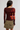A woman shown from the back wearing a long-sleeve maroon top with a large red and orange floral print on the back and sleeves, paired with an olive green mini skirt and a patterned belt, against a light gray studio background.