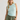 A woman wearing a light mint green sleeveless crop top, olive green pants, and a cream beanie with a gray floral graphic, standing against a light gray studio background.