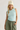 A woman wearing a light mint green sleeveless crop top, olive green pants, and a cream beanie with a gray floral graphic, standing against a light gray studio background.