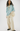 A model stands against a plain light gray studio background wearing a light blue knit sweater with a cream bow at the neckline, cream wide-leg pants, sheer ruffled sleeves, and black shoes.