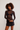 A model with curly brown hair wearing a sheer black mesh long-sleeve top over a black bralette, paired with a black denim mini skirt, against a plain light gray studio background.