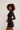 A woman with curly brown hair models a black long-sleeve mesh top and a dark denim mini skirt against a plain light gray studio background.
