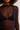 Close-up of a model wearing a black sheer long-sleeve top with a textured fishnet pattern over a black bralette, against a plain light background.