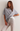 A woman wearing a loose, oversized heather gray t-shirt layered over a white undershirt and light beige jeans, posing against a plain off-white wall.