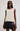 A woman modeling a sleeveless cream-colored knit vest with black faux-leather shorts against a plain light gray studio background.