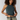 A woman with curly dark hair wearing a dark teal ribbed halter crop top and light olive pants with a patterned belt, posing against a light gray studio background.