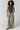 A model stands against a light gray studio background wearing a dark teal sleeveless crop top, wide-leg sage green pants, and black sandals.